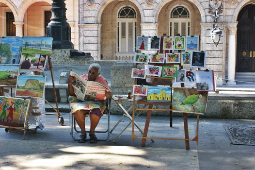 Ederly woman sitting,reading newspaper and selling pictures on the street in Havana