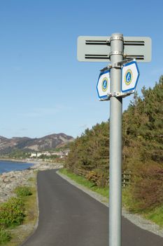 Signs for the 'Wales coast path'on a post with a tarmac track leading into the distance against a bright blue sky.