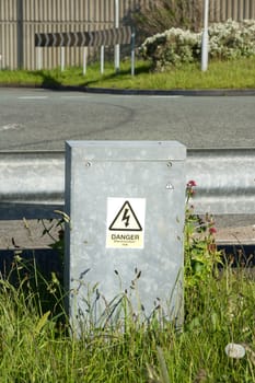 A sign for 'Danger' on a grey metal container in grass against a metal road barrier.