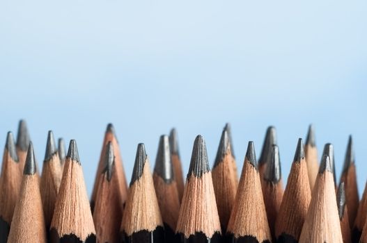 An uneven row of mixed graphite pencils, standing upright against a sky blue background with their tips pointing upwards from the bottom half of the frame.