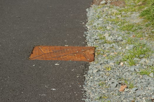 A tarmac road / path and edging hardcore with a rusty metal drain cover.