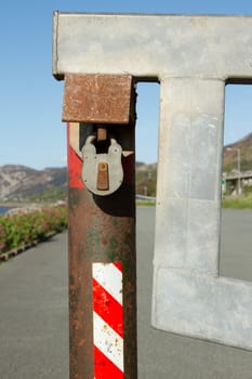 A metal gate, barrier attached to a rusty post with a padlock protected by a steel housing with a tarmac road in the background.