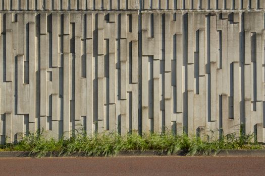 A precast concrete wall patterned with vertical oblong textures with green grass on a horizontal plane and a red tarmac path below.