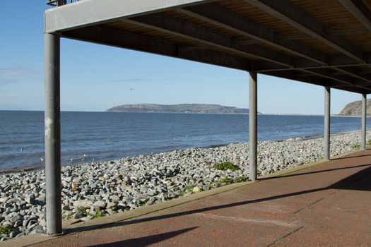 A path leads under a metal build veranda, coverway, with posts leading to a pebble beach and the sea and island beyond.