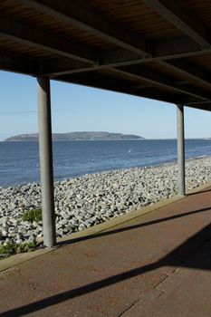 A path leads under a metal build veranda, coverway, with posts leading to a pebble beach and the sea and island beyond.