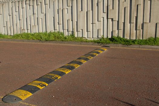 A black and yellow speed bump, rumble strip stretches across a red tarmac road.