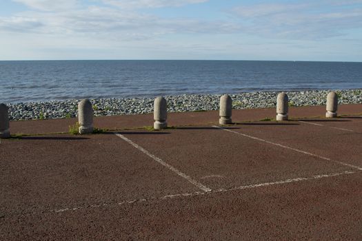 Concrete bollards at the edge of a car park with red tarmac and white lines, a pebble beach and the sea in the background.