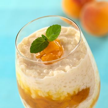 Fresh homemade rice pudding with peach compote garnished with mint leaf and served in a high glass (Selective Focus, Focus on the mint leaf and the front of the peach compote on top)