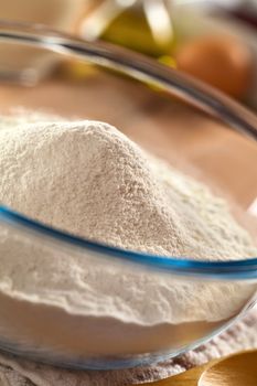 Preparing a batter (dough) for crepes or pancakes with wheat flour in glass bowl, and milk, eggs and oil in the back (Selective Focus, Focus on the top of the flour pile)