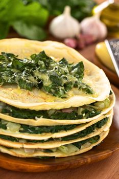 Fresh homemade savory crepes layered with chard (mangold) and onion with cheese on top served on wooden plate with ingredients in the back (Selective Focus, Focus one third onto the top of the crepes) 