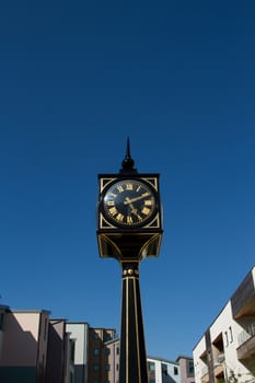 A clock with roman numerals on a post, pillar, painted black and gold with generic housing and a blue sky as a background.