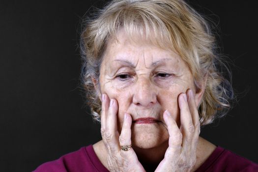 Dramatic portrait of a sad, depressed or worried senior woman with hands in her face, studio shot over black background.