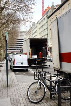 Typical busy city related or urban scene with delivery or moving trucks, parked cars, bicycle and large buildings.