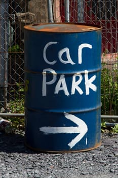 An oil drum painted blue with a sign painted in white with the words 'CAR PARK' with an arrow pointing left.