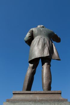 The rear view of a statue cast in bronze on a slate plinth with a man gesticulating in a frock coat against a clear blue sky.