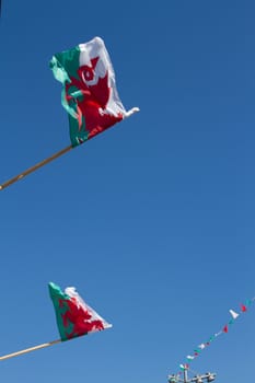 A pair of Welsh flags on poles fluttering and bunting in the distance against a clear blue sky.