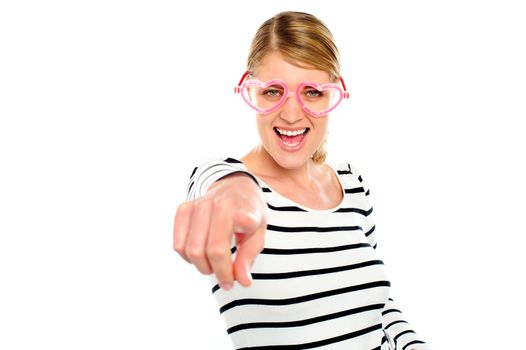 Excited woman wearing heart shaped eye-wear pointing at you isolated over white background