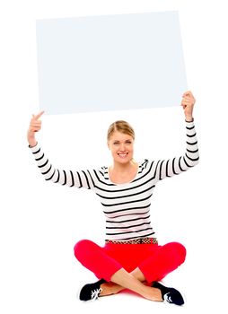 Beautiful young woman sitting on floor pointing towards blank banner ad over her head