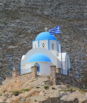 Small blue domed church suurounded with a fence and on a litlle hill next to a greek flag at Kamari, Santorini, Greece