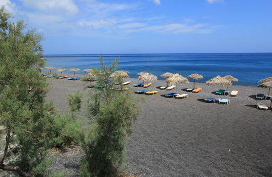 Black beach with umbrellas made of straw and colorful deckchairs at Kamari, Santorini, Greece, by beautiful weather.