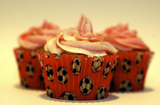 Three cupcakes with pink frosting on the stop, in red cases decorated with tiny footballs, against a white background with copy space.