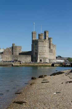 The historic castle in the town of Caernarfon, Gwynedd, Wales, UK, with the Menai Strait in the foreground and a blue sky in the background.