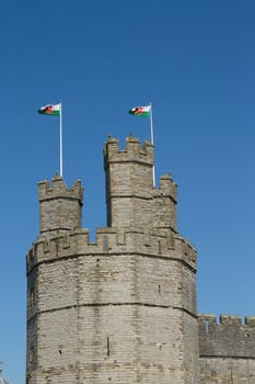 The towers, walls and ramparts of a castle with two Welsh flags flying against a clear blue sky.