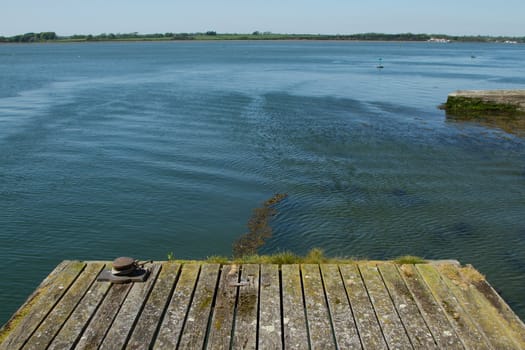 An old wooden dock with lichen and weed and a metal mooring point with a rope with the sea and distant island fields.