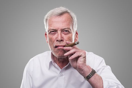 Handsome senior man with a shrewd expression and strong personaity smoking a cigar isolated on a grey studio background