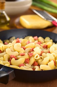 Preparing fresh homemade elbow macaroni pasta with sausage pieces in frying pan with cheese, oil and green onion in the back (Selective Focus, Focus one third into the frying pan)