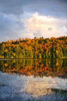Forest of colorful autumn trees reflecting in lake with dramatic sky