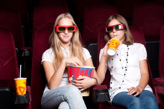 two girls look three-dimensional cinema, sitting in the glasses, eat popcorn, drink drink