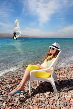 Young girl in sunglasses and a white hat resting on the sea, windsurfer in the background