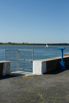 A blue telescope placed by a quay wall with metal gates with a yacht in the sea and fields in the distance.