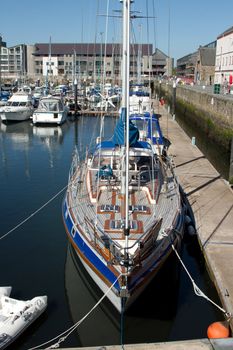 A yacht moored in a marina with a footpath access below the dock walls with buildings in the background and a blue sky.