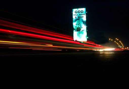 Religious signboard at night in a  busy expressway with streak of colorful lights caused by passing vehicles' rear and back lights.