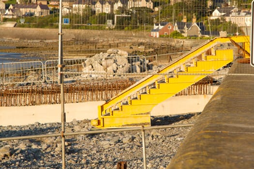 A set of steel temporary steps painted yellow leads to a construction site on a pebble beach.