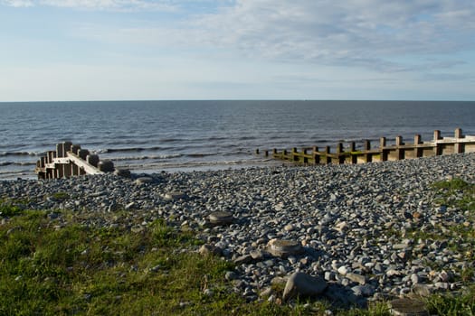 A pebble beach with coastal protection barriers made of wood with the sea and blue cloud sky in the distance.