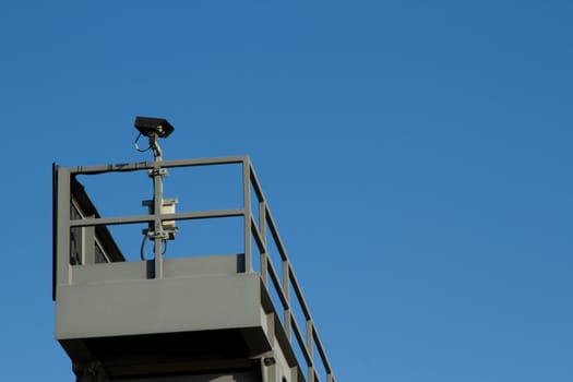 A security camera placed on a metal stanchion with walkway against a clear blue sky.