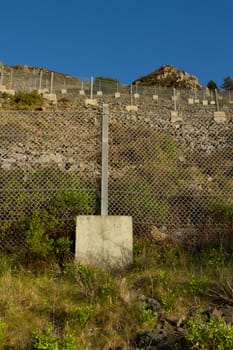 An area of steep loose rocky ground with a series of safety, retaining, fences with linked metal wire, metal posts and concrete supporting blocks,