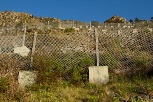 An area of steep loose rocky ground with a series of safety, retaining, fences with linked metal wire, metal posts and concrete supporting blocks,