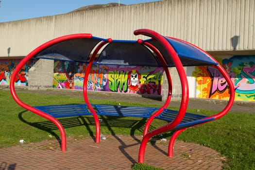 An unusual park seat on bricks with green grass and a graffiti wall in the background.