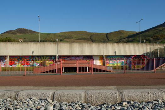 A pavement in front of a fenced skate park with ramps and a graffiti wall behind with hills and a clear blue sky in the distance.