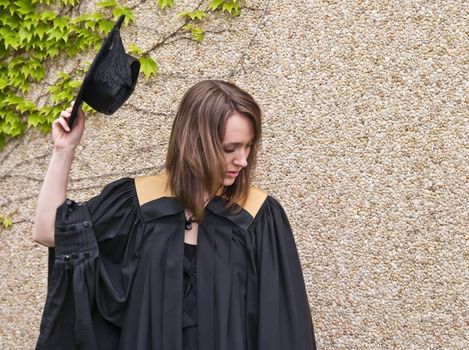 Young woman holding graduation hat against stone and leafy wall.