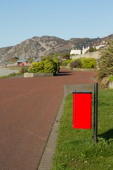 A red bin on a post for dog poo, faeces, next to a footpath with green grass borders and a hill in the distance.