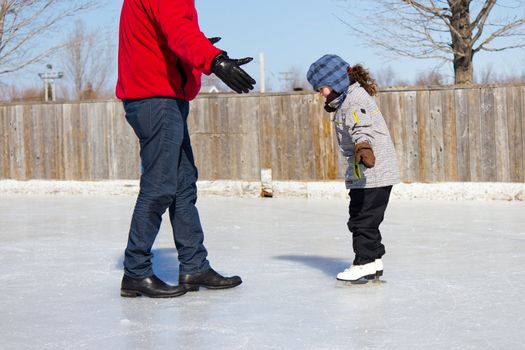 Father teaching daughter how to ice skate at an outdoor skating rink in winter.