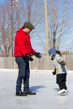 Father teaching daughter how to ice skate at an outdoor skating rink in winter.