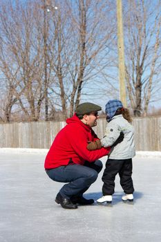 Father teaching daughter how to ice skate at an outdoor skating rink in winter.