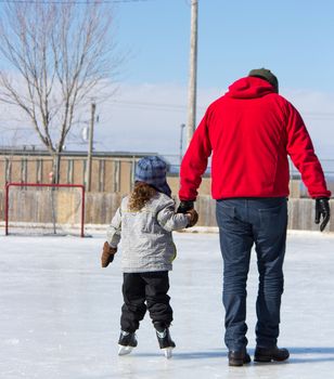 Father teaching daughter how to ice skate at an outdoor skating rink in winter.