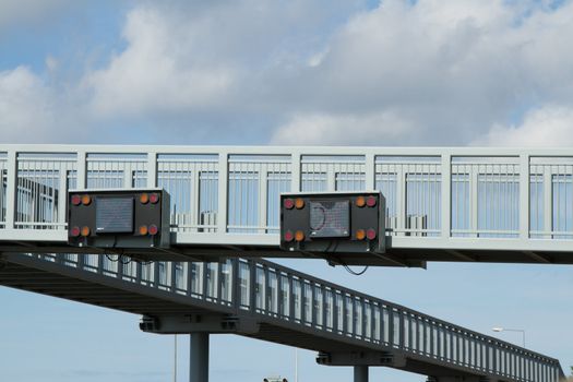 A pair of electronic traffic signs fixed to a raised metal walkway with a cloudy sky in the background.